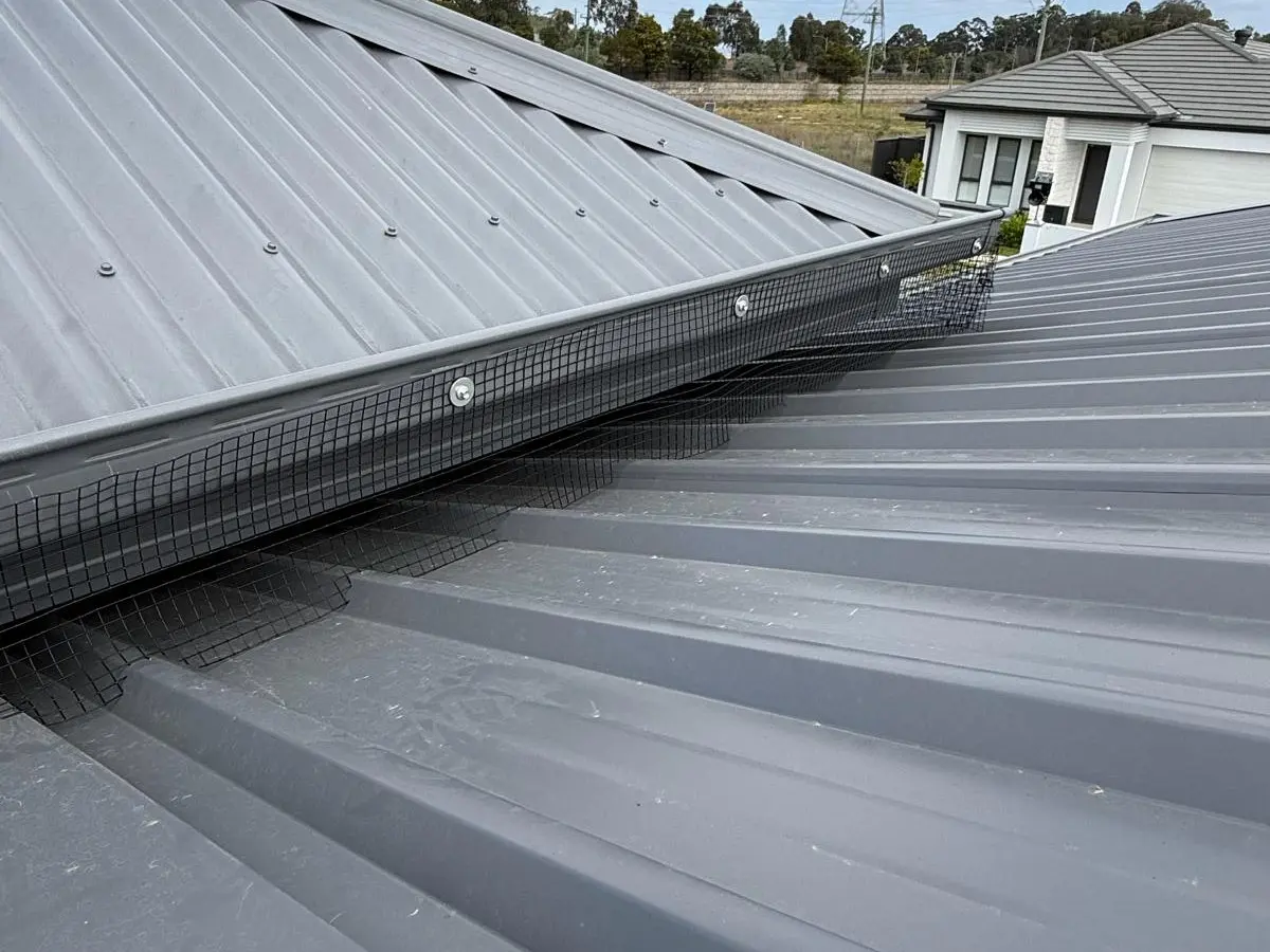 Black bird-proofing mesh fixed along the join where an upper metal roof steps down onto a lower one, on a suburban house.
