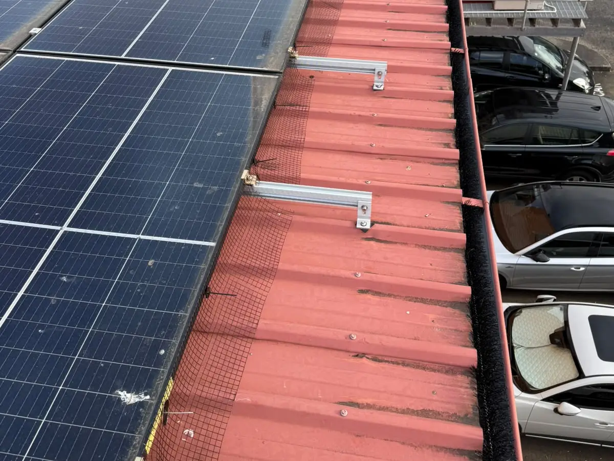 Mesh fitted around the edge of a solar array on a red corrugated metal roof, with a black gutter brush in the gutter at the roof edge.