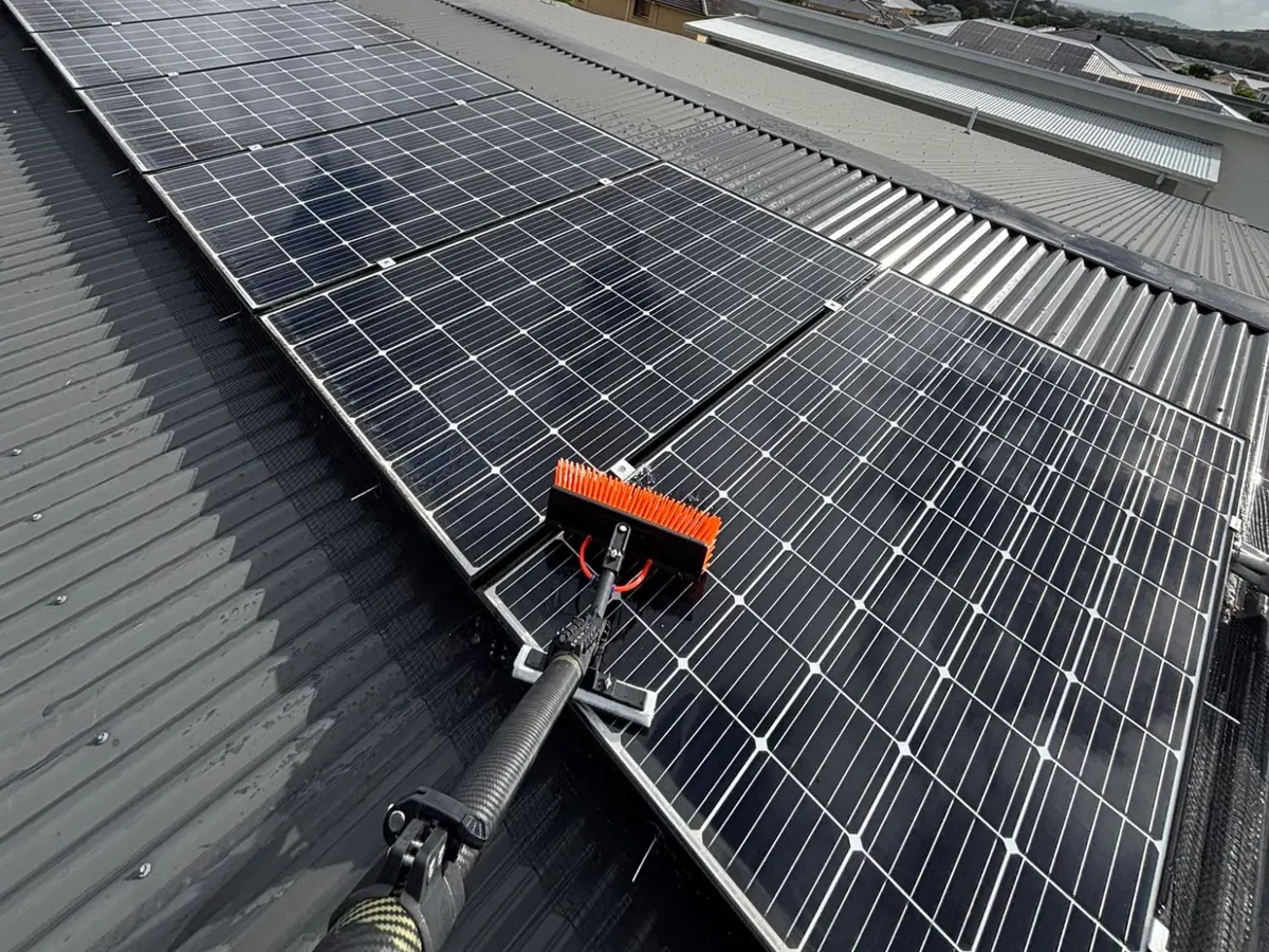 A soft orange brush on a carbon extension pole cleaning a wet rooftop solar array on a dark metal roof, with mesh along the edge of the panels.